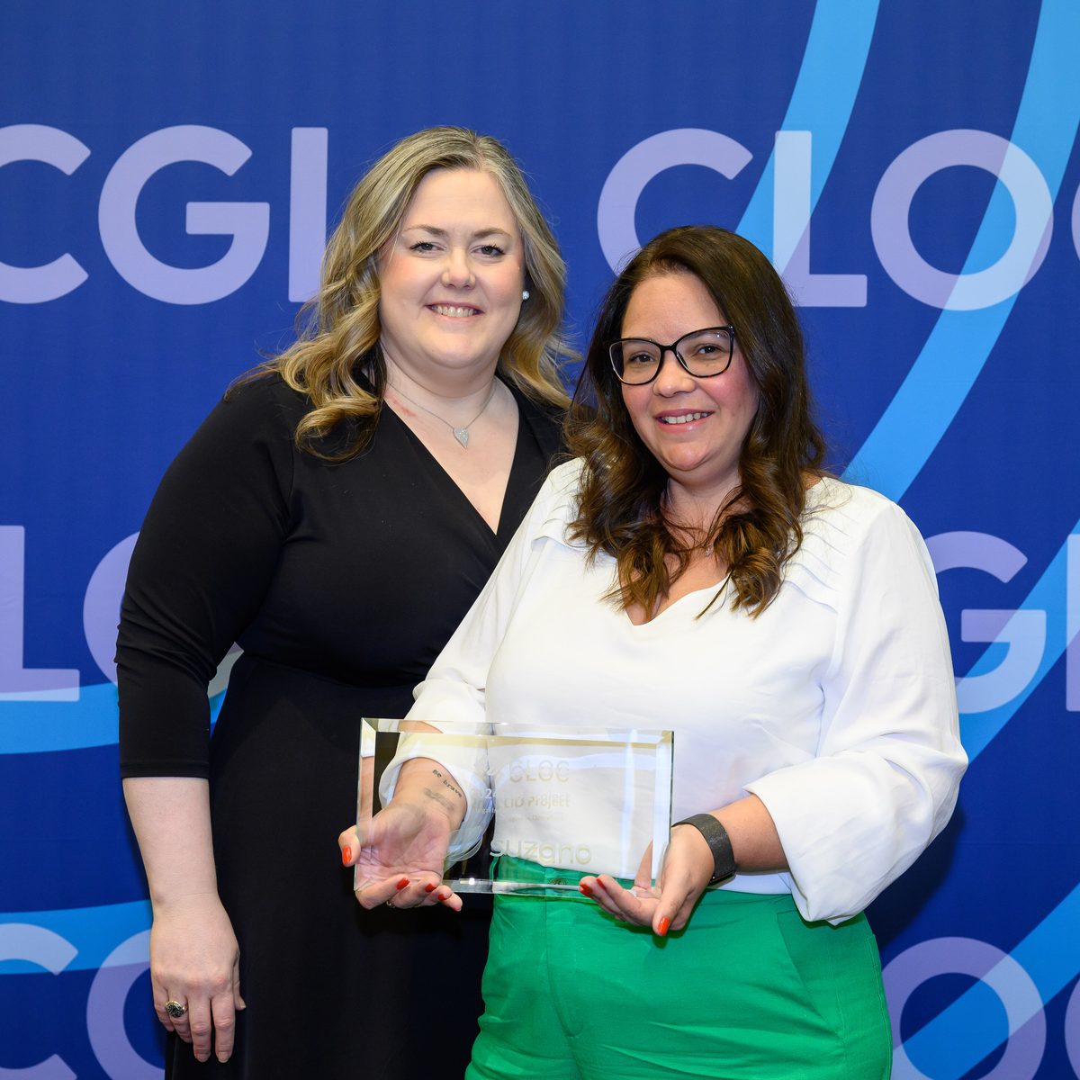 Two women smile in front of a blue CLOC CGI backdrop as one holds a glass CLOC award